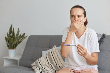 Beyaz tişört giyen hamile bir kadın elinde hamilelik testi, evinde koltukta oturuyor, ağzını ellerle kapatıyor, kötü hissediyor, zehirlenmiş ve bulantısı var..