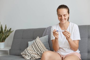 Gülümseyen, iyimser, koyu saçlı, beyaz giyimli, hamilelik testi sonucu pozitif çıkan, oturma odasında otururken poz veren, beyaz saçlı kadın..