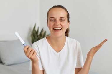 Son derece mutlu, hamile bir kadın, yatak odasında hamilelik testi pozu veriyor, beyaz tişört giyiyor, elini kaldırıyor, neşeyle neşe içinde kameraya bakıyor..