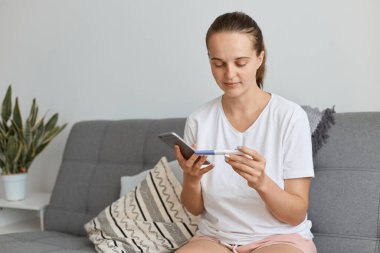 İyimser hamile, beyaz saçlı, günlük kıyafet giyen, elinde hamilelik testi olan, elinde akıllı bir telefon tutan, iyi haberleri vermek için koca numarasını arayan bir kadın..