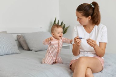 İç mekanda, elinde at kuyruğu olan, elinde hamilelik testi olan, küçük kızıyla yatak odasında oturan, bebeğine bakan, sonucunu bekleyen çekici bir kadın fotoğrafı..