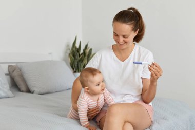 Hamile bir kadının bebek bebekle yatak odasında poz verirken, kızına sarılırken, tekrar anne olmaktan mutlu olurken, beyaz t-shirt giyerken..
