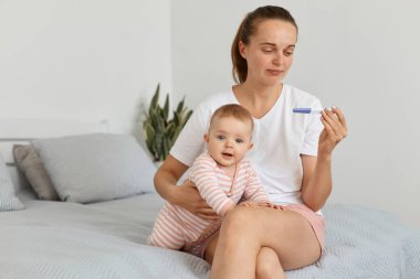 Mutsuz, hamile ve hamile bir kadının beyaz t-shirt 'ü içinde, bebek bebekle yatak odasında poz verirken, pozitif sonuç görmek için üzüntüsünü ifade ederken..