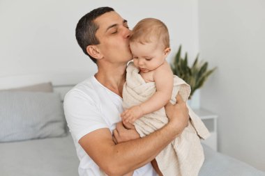Kapalı alanda esmer bir adam, beyaz bir tişört giyen sevgili bir baba küçük kızını havluya sarmış, sevgi ve özenle başını öpüyor, aydınlık odada poz veriyor..
