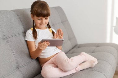 Şirin gülümseyen, at kuyruklu, hafif oturma odasındaki koltukta oturan, elinde telefon olan, video konuşması yapan ya da çocuklar için canlı yayın yapan sevimli bir kız..