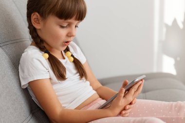 Evdeki koltukta oturan, arkadaşlarıyla iletişim kurmak için cep telefonu kullanan ya da sosyal ağlardaki çocukları kontrol eden beyaz tişörtlü sakin küçük kızın yan görünümü..