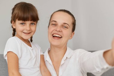Güzel kızıyla selfie çeken, cep telefonu kamerasına gülümseyen, mutluluğu ifade eden mutlu bir kadının kapalı mekan fotoğrafı, evde poz veren bir aile fotoğrafı..