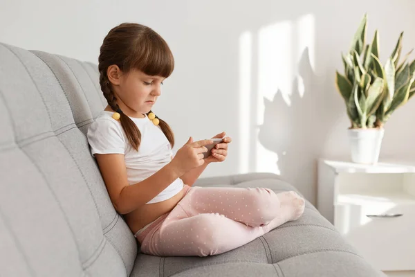 聚精会神地坐在沙发上 玩电子游戏 穿着白色休闲衫的可爱的深色头发女婴的侧视图肖像 — 图库照片