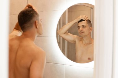 Banyoda aynanın önünde duran çıplak vücutlu yakışıklı adam, sabah duş aldıktan sonra poz veriyor, aynadaki yansımasına bakıyor ve saçına dokunuyor..