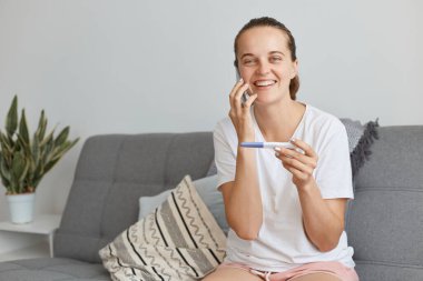 Son derece mutlu, siyah saçlı, beyaz tişörtlü, kısa boylu, oturma odasında koltukta oturan ve telefonla konuşan, hamileliğini olumlu bir testle anlatan bir kadın..