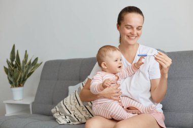 Gülümseyen genç anne yeni yürümeye başlayan bebeğiyle yatakta oturuyor ve hamilelik testi pozitif sonuç veriyor, tekrar anne olmaktan tatmin oluyor, mutluluğu ifade ediyor..