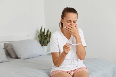 Hamile, çekici bir kadın, gündelik kıyafetlerini giyip evdeki kanepede oturuyor ve hamilelik testi pozitif sonuç veriyor. Zehirlenme ve mide bulantısı var..
