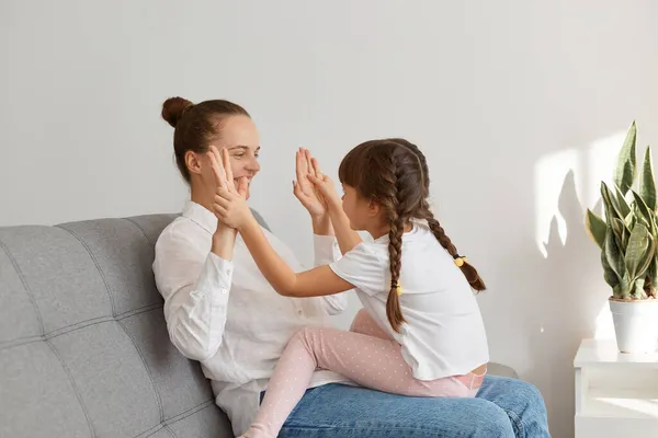 Indoor shot of happy mother playing with her charming daughter with pigtails pet a cake, expressing positive emotions, sitting together on gray sofa in living room.
