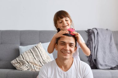 Gülümseyen, çekici bir adam ve küçük kızının portresi, aile zamanı ve birlikte eğlenmek, kanepenin yanında oturmak, kızın babasına komik saç stili yapması, olumlu duygular ifade etmesi..