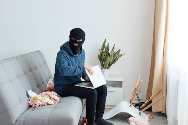 Kapalı alanda, koyu mavi kapüşonlu ve siyah maskeli bir erkek soyguncunun başkasının evine girip bir dizüstü bilgisayar çalmaya çalışırken, kanepede oturur ve kameraya bakar..