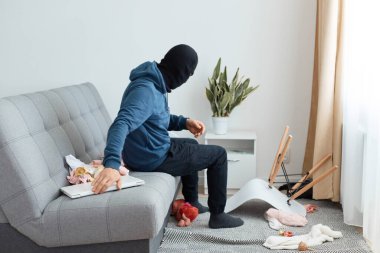 Hırsız evde soymaya çalışan bir laptop bulmuş, kanepeye oturmuş, komodini kontrol ediyor, çalacak başka bir şey arıyor, mavi kapüşonlu ve siyah kar maskeli bir adam soyguncusu.