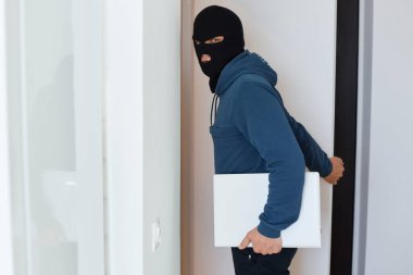 Soyulan bir evde mavi kapüşonlu ve siyah kar maskeli bir erkek soyguncu, kameraya bakıyor, elinde çalıntı bir dizüstü bilgisayar tutuyor, bir erkek yasadışı eylemlerde bulunuyor..