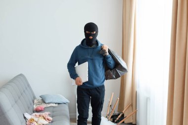 Davetsiz misafir kanepe ve pencerenin yanında duruyor ve elinde çalıntı eşyalar ve dizüstü bilgisayarla büyük bir çanta tutuyor. Kameraya bakıyor. Mavi kapüşonlu ve siyah kar maskeli bir adam..