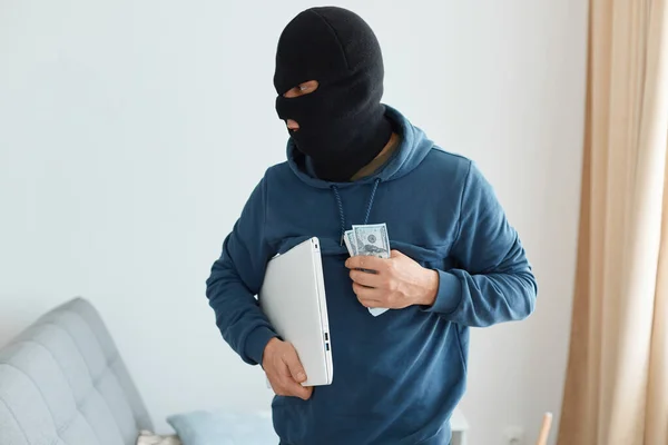 入室盗窃的男子身穿深蓝色帽衫 戴着黑色面具 手持手提电脑 在别人家里偷了一大笔钱 离开了非法行动的地方 — 图库照片