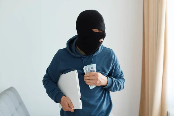 男子抢劫犯身穿蓝色帽衫和黑色巴拉克拉瓦抢劫手提电脑 在别人的软管里赚钱 离开犯罪地点 看着窗户 非法行动的画像 — 图库照片
