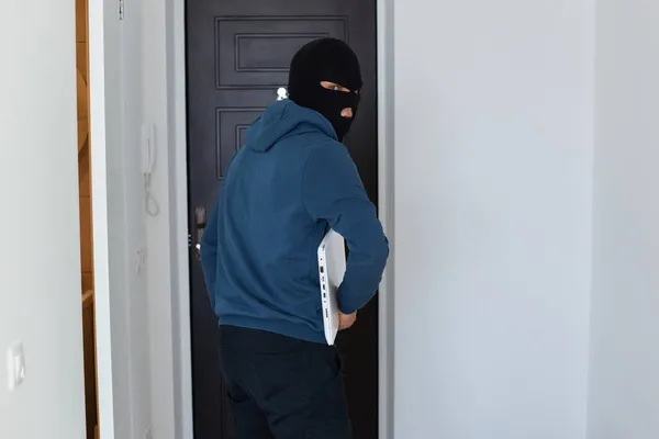 身穿深蓝色帽衫 戴着黑色面具 手里拿着偷来的手提电脑 偷了别人的东西 被吓坏了的男性窃贼离开了他抢劫的房子 — 图库照片