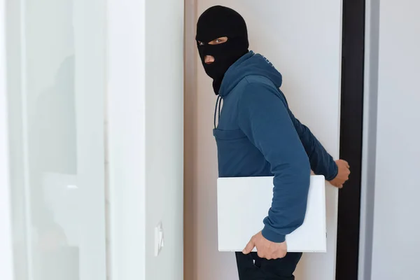 一名身穿蓝色帽衫 头戴黑色巴拉克拉瓦的男子在抢劫的房子里摆姿势 看着摄像机 手持被盗的笔记本电脑 犯了非法行为 — 图库照片