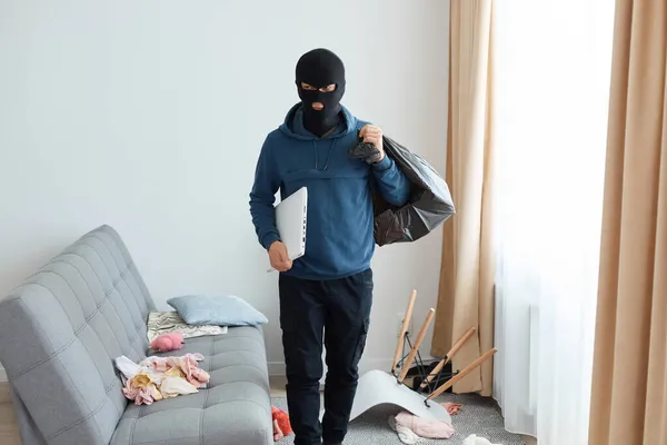 男子抢劫犯身穿蓝色帽衫 头戴黑色巴拉克拉瓦 站在沙发和窗前 手里拿着装有被盗物品和手提电脑的大包 望着别处 找到出口的画像 — 图库照片