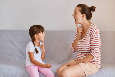 Profilde kadın fizyoterapistin doğru konuşma için küçük kız çocuğundan ders aldığı, insanların kanepeye oturduğu, küçük çocukların konuşmalarını geliştirdiği..