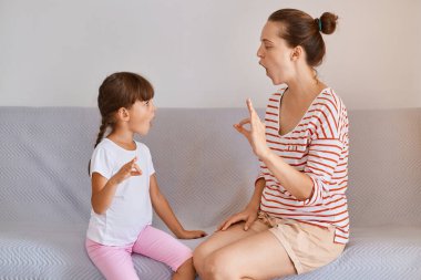 Kanepede otururken küçük bir kızla poz veren kadın fizyoterapistin kapalı alandaki görüntüsü konuşma sorunlarını çözer, sesleri nasıl telaffuz edeceğini gösterir..