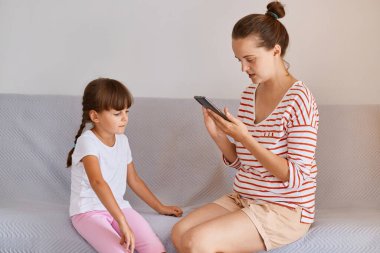 Küçük bir kızla çalışan, elinde cep telefonuyla koltukta oturan profesyonel kadın fizyoterapist, eğitim için aletinde yeni bir online ders buluyor..