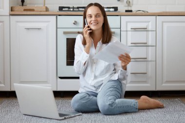 Kot pantolon ve gömlek giyen, beyaz mutfak tezgahında oturan, akıllı telefonla konuşan, elinde belgelerle dizüstü bilgisayarın yanında poz veren, iyimser, beyaz saçlı, iyimser bir kadın..