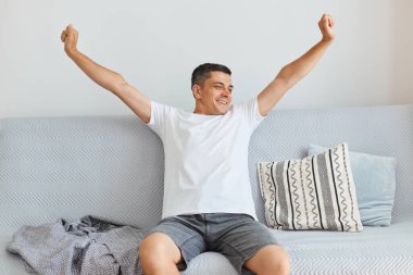 Beyaz gömlek ve kot giyen, açık oturma odasındaki gri koltukta oturan, kollarını açarak uyanan, mutlu ve çekici esmer adamın kapalı çekimleri..