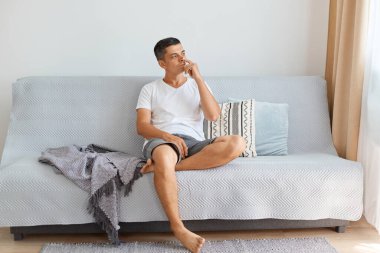 Beyaz gömlek ve kot pantolon giyen komik, düşünceli genç bir adamın portresi, düşünceli bir yüz ifadesiyle başka yere bakıyor, parmağını burnuna sokuyor, kanepede otururken içeride poz veriyor..