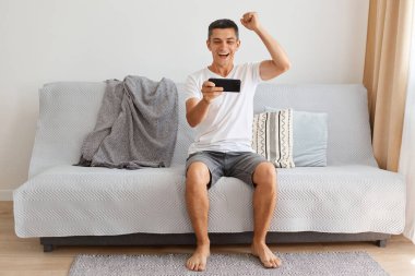 Heyecanlı, siyah saçlı, beyaz tişörtlü, kısa pantolonlu, akıllı telefonla video oyunu oynayan, yumruğunu sıkan, galibiyeti kutlayan, rahat bir koltukta oturan genç bir erkek..