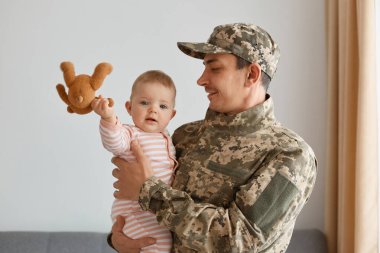 Kapalı alanda gülümseyen asker üniforması ve şapkası giymiş küçük kızıyla elinde oyuncak bebekle durup babasıyla oynadığı için mutlu olduğu bir fotoğraf..