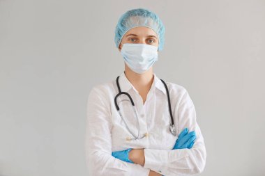 Eldiven, tıbbi maske ve kep takan doktor portresi, kolları çapraz kapalı poz veren, kendine güvenen yüz ifadesine sahip, hastaları bekleyen....
