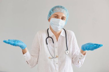 Kafası karışık doktor portresi eldiven, tıbbi maske ve kep takan, önlük giyen, kollarını açıp kameraya bakan, boynunda stetoskop olan.