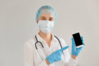 Tıbbi başlık, eldiven, ameliyat maskesi ve cübbesi takan pozitif kadın doktor, reklam veya promosyon için boş ekranlı cep telefonunu gösterip duruyor..