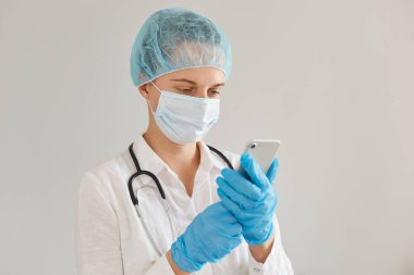 Kapalı alanda, tıbbi başlık, eldiven, ameliyat maskesi ve cübbesi takan, ayakta duran ve cep telefonu kullanan, e-postaları kontrol eden ya da birini arayan kadın doktorların fotoğrafları..