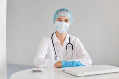 Kapalı alanda, tıbbi başlık, ameliyat maskesi, lastik eldiven ve önlük takan, çalışmak için not defteri kullanan, masada otururken kameraya bakan kendine güvenen kadın doktorların görüntüleri..
