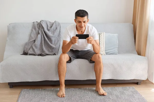 全神贯注的白人黑发男子 身穿白色T恤 身材矮小 坐在灰色沙发上摆出轻松的姿势 通过手机玩游戏 表情镇定 — 图库照片