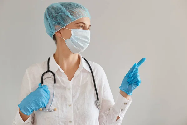 女医生头戴医用帽 外科口罩和长袍 站在那里用食指指尖指尖 展示广告复制空间的画像 — 图库照片