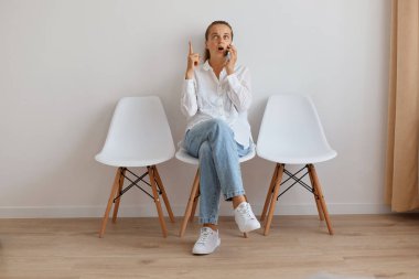 Süslü kot pantolon ve beyaz gömlek giyen, cep telefonuyla konuşan, parmağıyla işaret eden, ışık duvarına yaslanmış at kuyruklu, genç ve çekici bir kadın..