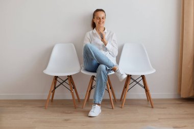 Beyaz gömlek ve kot pantolon giyen gülümseyen yetişkin beyaz kadın portresi sandalyede oturuyor ve akıllı telefon tutuyor, başka tarafa bakıyor, mutlu bir yüz ifadesi takınıyor..