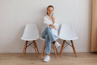 Beyaz gömlek ve kot pantolon giyen, sosyal ağları kontrol etmek ya da mesaj yazmak için akıllı telefon kullanan, pozitif gülümseyen beyaz kadın..