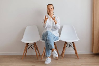 İyimser, çekici, at kuyruklu, ışık duvarına yaslanmış sandalyede oturan, şık kot pantolon ve beyaz gömlek giyen, kamerayı işaret eden kadının tam boy portresi..