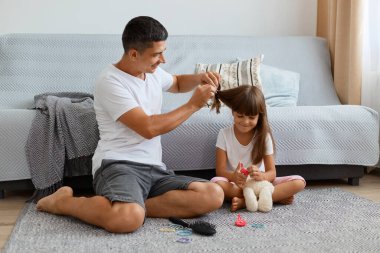 Mutlu, esmer, beyaz tişört ve kot pantolon giyen yakışıklı erkek. Kızıyla birlikte yerde oturuyor ve at kuyruğu yapıyor. Kanepenin yanında poz veriyor..