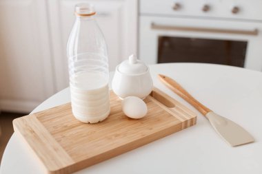 Yemek pişirmek için kapalı alanda çekilen ürünler, şişede süt, şeker, yumurta, kesim tahtası, şeker, mutfak spatulası, arka planda beyaz mutfak setiyle masada duran.