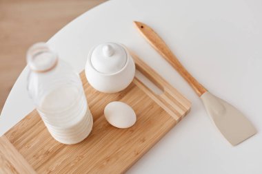 Şişedeki sütün üst görüntüsü, şeker, yumurta, kesim tahtası, şeker, mutfak spatulası, beyaz masada duran, pişirme malzemeleri..