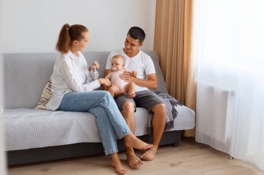 Romantik genç çift gündelik giysiler giyip çocuklarıyla vakit geçiriyorlar. Küçük bir bebekle, ebeveynlik yaparak, açık salonda kanepede oturuyorlar..